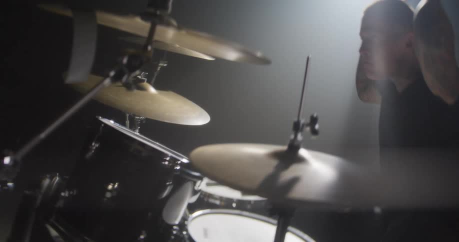 The camera pans vertically from the polished concrete floor and chrome hardware to reveal a tattooed drummer fiercely striking his kit under a hazy spotlight
