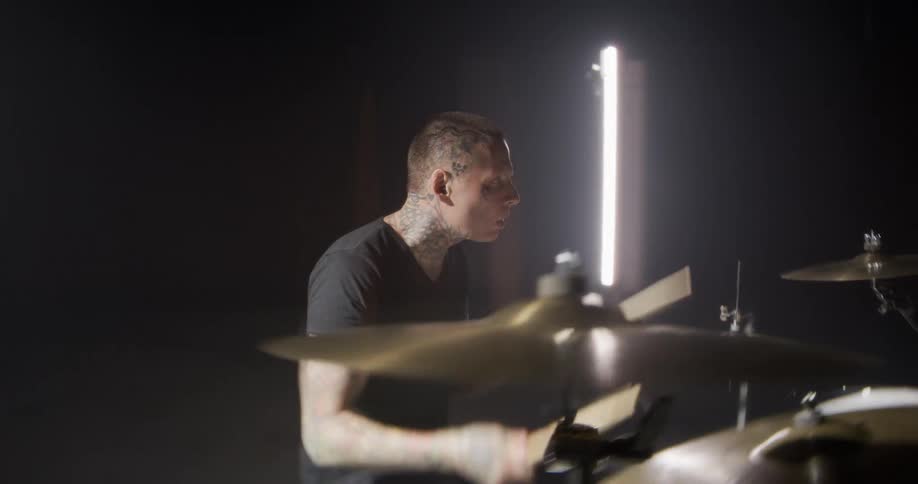A man with tattoos plays a drum set energetically under bright lights in a dark room