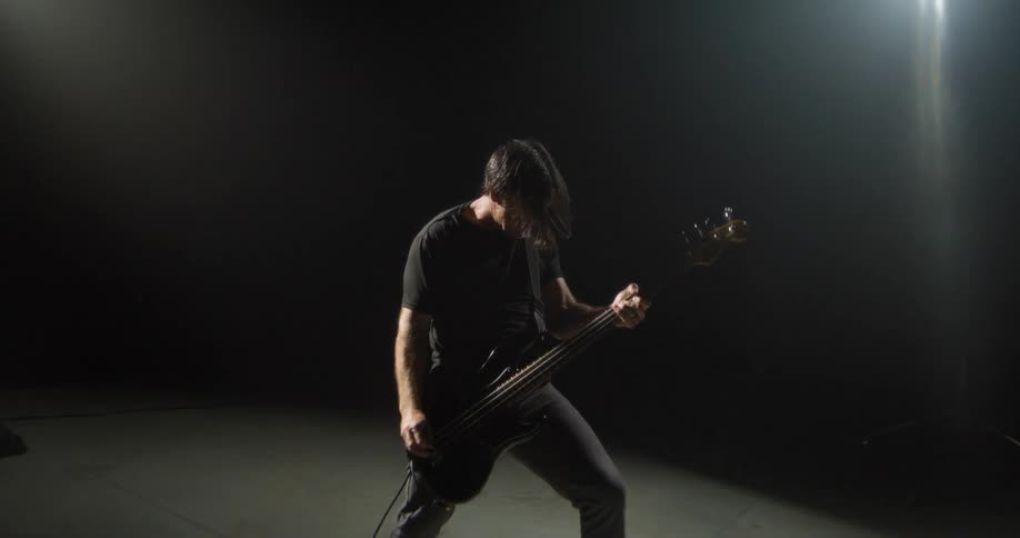 A man with long dark hair plays a bass guitar energetically in a dark room under a single bright spotlight