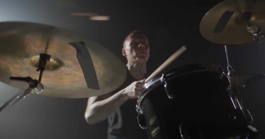 A tattooed drummer in a black tshirt performs an intense set under moody cinematic lighting the camera pans around the drum kit as he strikes the cymbals and snare with rapid precision