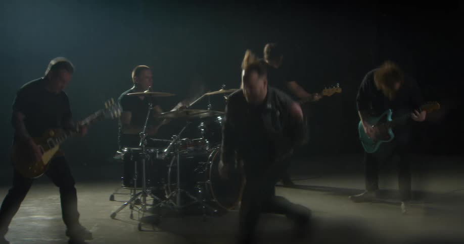 Band performing in a dark studio with a lead vocalist at the center guitarists bassist and drummer under directional lighting