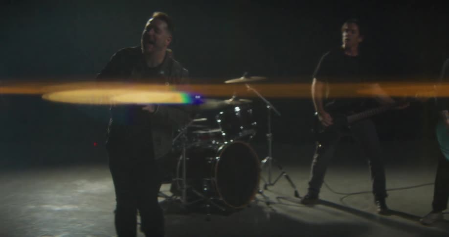 Band performing in a dark studio with a lead singer in the foreground guitarists and drummer in the background under directional lighting