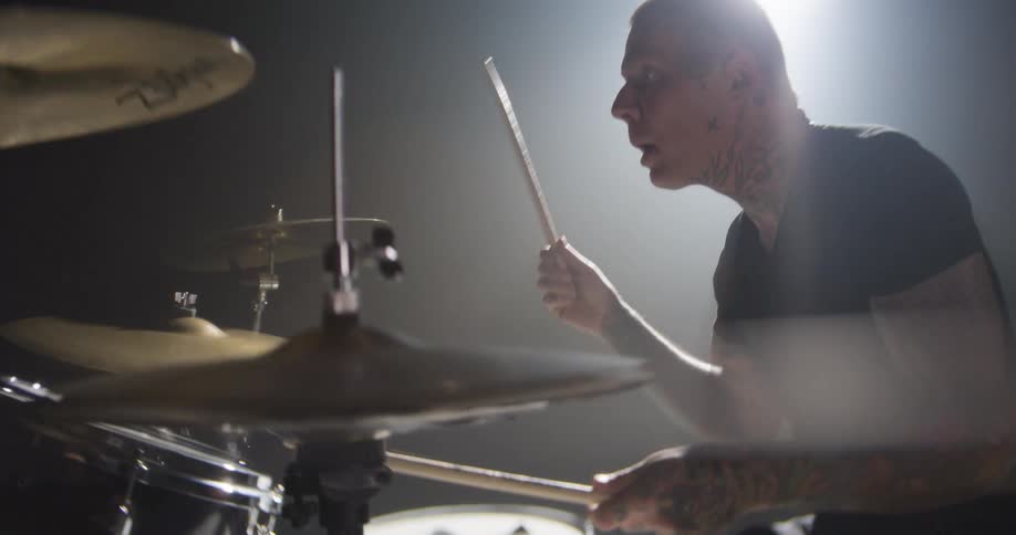 A heavily tattooed drummer energetically plays a drum kit under a bright spotlight