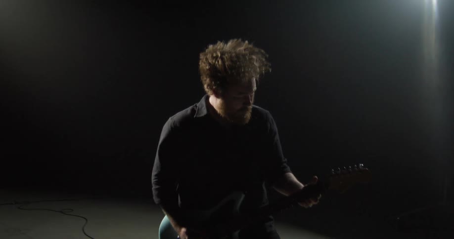 A bearded man in a dark studio vigorously plays a light blue electric guitar while head banging under a bright spotlight