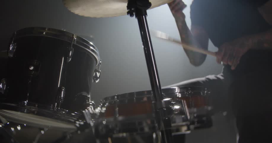 A drummer with tattooed arms plays a black and orange drum kit in a dimly lit smoky roomthe camera provides a closeup lowangle view as the musician strikes the cymbals and drums with wooden sticks