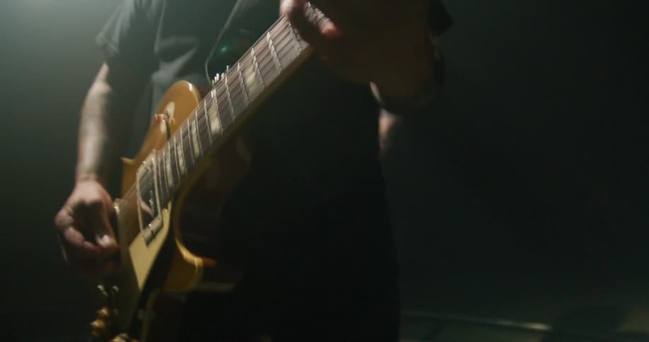 The video shows a closeup of a musician playing an electric guitar in a dark atmospheric setting a bright spotlight shines from behind the performer creating a heavy backlit effect and casting much of the scene in shadow