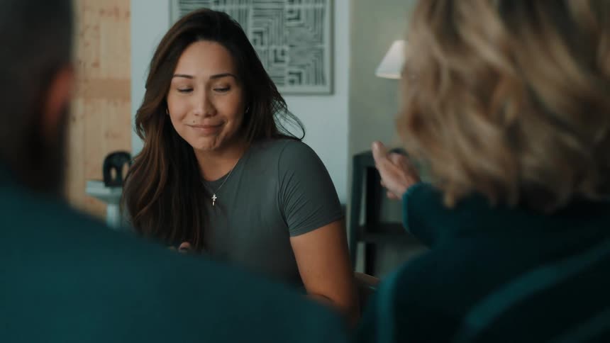 A woman in a grey shirt shares a warm smile while listening to a conversation with her family around a table
