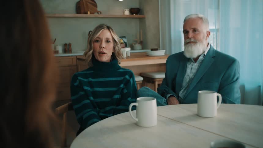 Two adults seated at a table in a home setting with mugs in front and a kitchen in the background