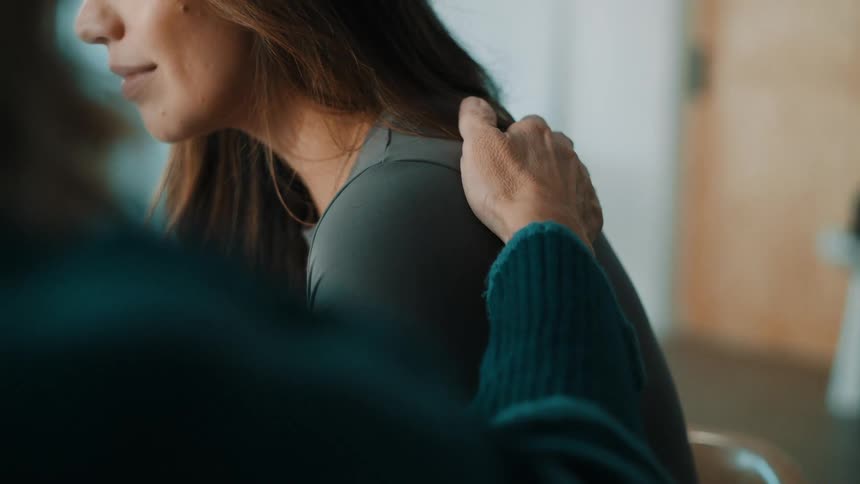 A close view shows a person placing a hand on a womans shoulder as a supportive gesture while they sit together