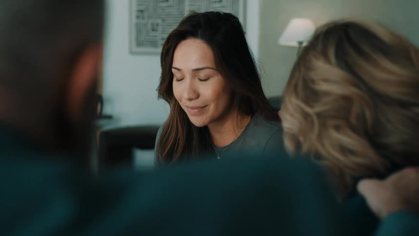 A woman sits with her eyes closed and a gentle smile appearing to pray while gathered with two other people whose backs are to the camera in a dimly lit modern living room