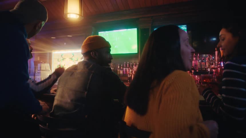 Black adult man wearing a yellow beanie sitting on the counter of a bar chatting with adult women around him indoors during night time