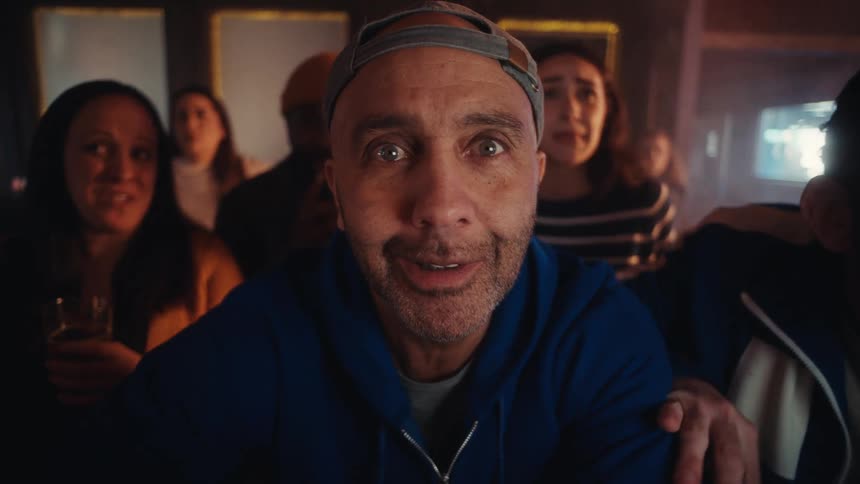 Man staring directly into a bar setting with other people reacting in the background under warm lighting