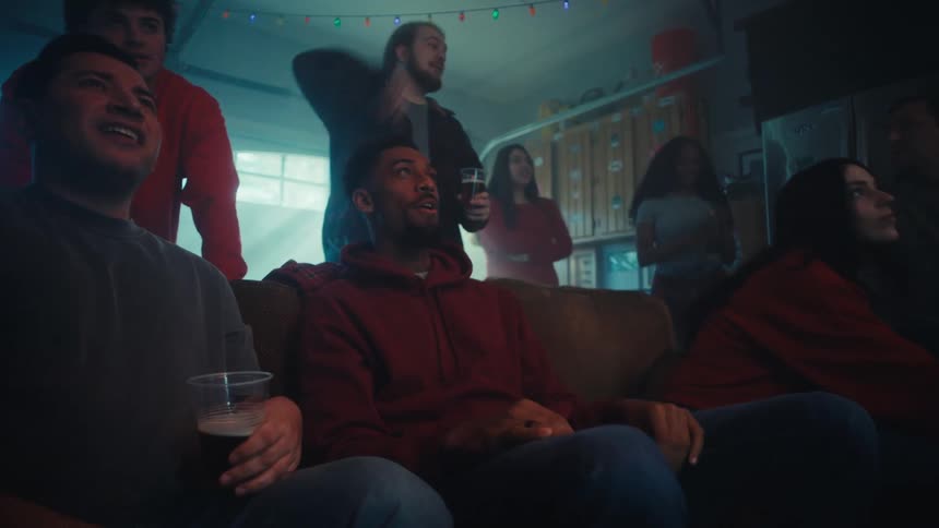 A group of friends sits on a couch in a dimly lit room reacting with intense focus and excitement while watching an event on television