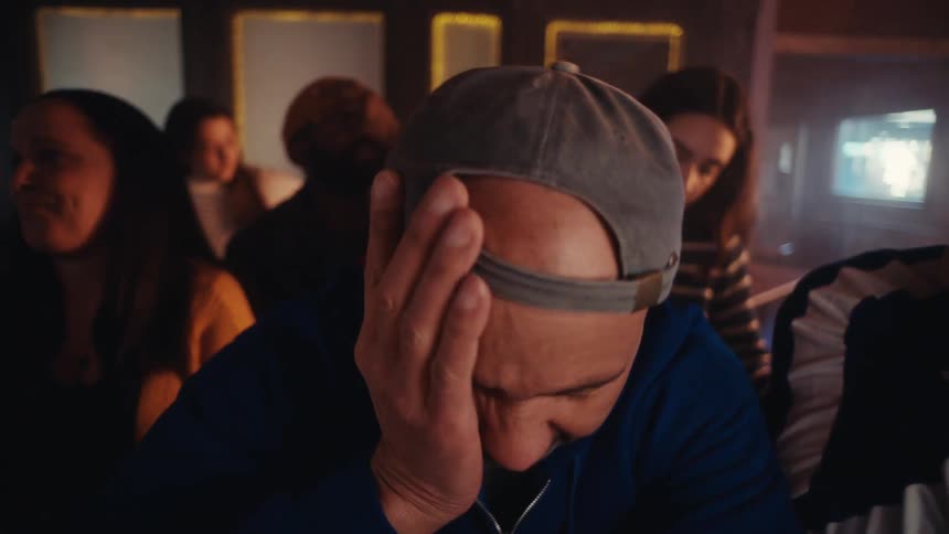 A man wearing a blue hoodie and a grey baseball cap looks distressed and covers his face with his hand while a crowd of people stands behind him in a dimly lit room