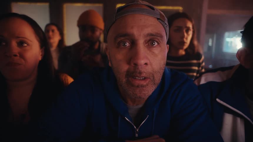 Group of people in a bar watching something offscreen with a man in the foreground reacting under warm lighting