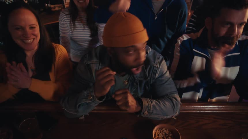 Young adult black man wearing an orange beanie screaming and celebrating at a bar counter surrounded by also people also cheering and clapping indoors