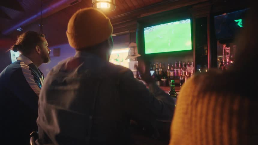 Group of people seated in a bar watch a soccer match broadcast on television