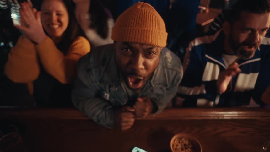 Young adult black man wearing a beanie screaming and cheering at a bar counter surrounded by other people also cheering and clapping indoors