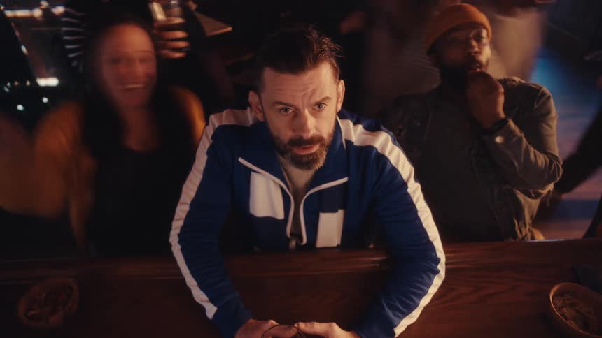 A man is sitting alone at a bar as other people talk and laugh behind him