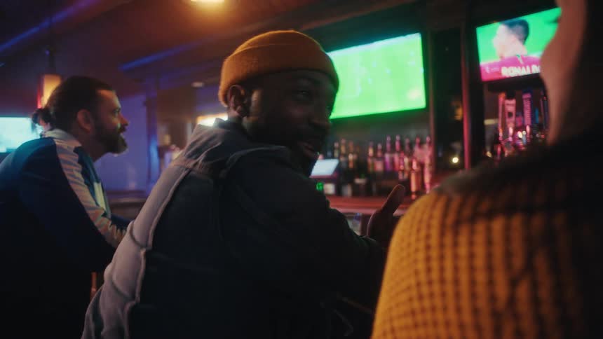 A man in a denim jacket and a yellow beanie cheers while watching a soccer game with friends at a sports bar