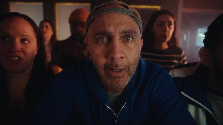 A close view shows a middle aged man wearing a blue hoodie and a backwards baseball cap staring intently with an expression of shock and disbelief