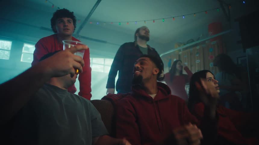 A group of friends sitting in the living room watching a game with excitement some are holding cups of beer and they suddenly feel disappointed by the result