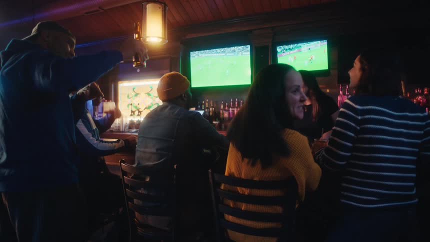 People seated and standing around a bar counter following a sports broadcast on televisions in a bar setting