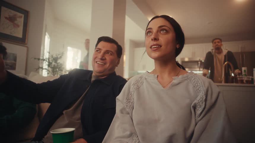Couple seated in a welllit living room looks at something offscreen while other people remain in the background