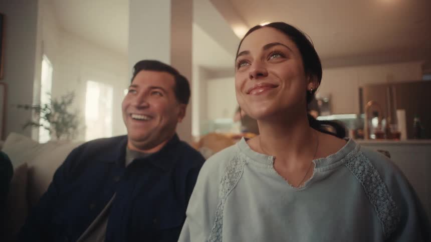 A woman in a blue sweatshirt and a man in a navy jacket sit together on a sofa looking toward an offscreen television while smiling and laughing in a dimly lit modern living room with a kitchen visible in the background