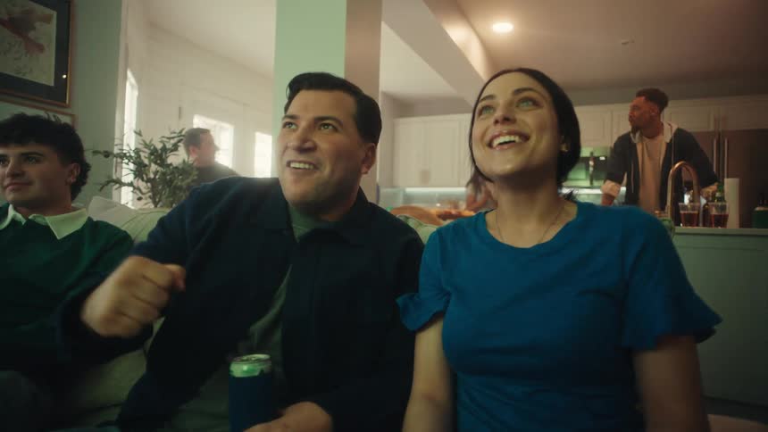A group of friends sits on a couch in a brightly lit home reacting with excitement and cheers while watching a screenthe man and woman in the foreground laugh and gesture enthusiastically as they celebrate a moment in the program