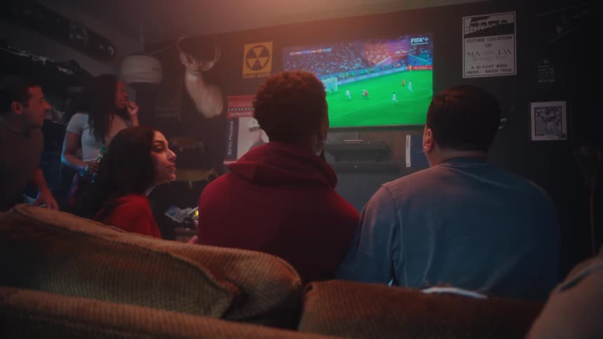 A group of friends watches a soccer match on television together in a dimly lit room