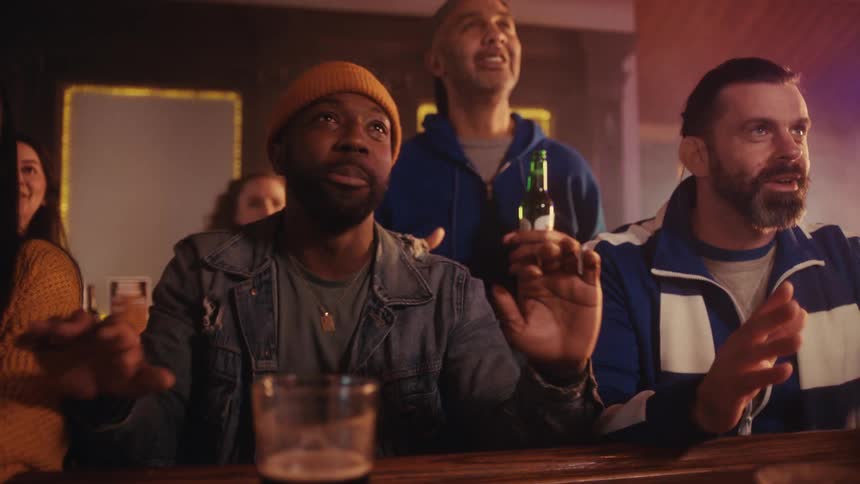 People in a bar reacting to an offscreen broadcast with hand gestures and drinks on the counter