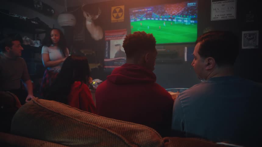 Group of young friends from different etchnicities talking and eating snacks while watching a soccer game on a television indoors