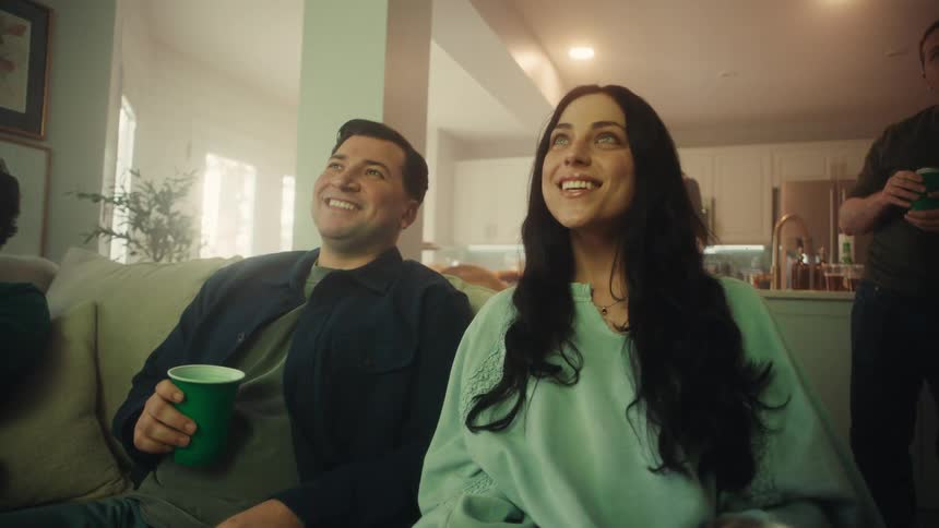 A group of friends sits together in a living room and reacts with wide smiles and cheers while watching an event on television