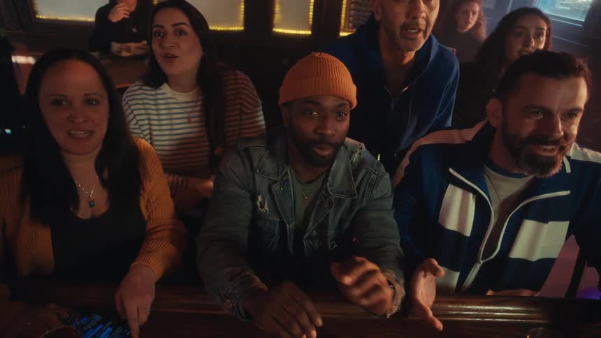 People sitting and standing at a bar talking and looking up at the television