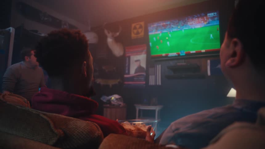 In a dimly lit room a group of friends sits on a couch while intently watching a soccer match on a large television screen the atmosphere is casual and focused with one person holding a bowl of snacks as they react to the action on the field