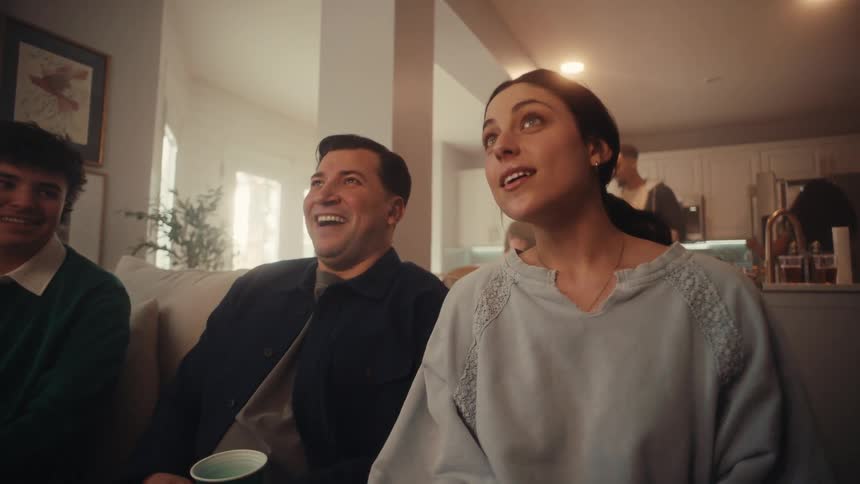 A group of friends sits on a sofa looking upward with focused and expectant expressions as if watching a game or eventas the video progresses their tension turns into visible joy and excitement with the man in the center breaking into a wide happy