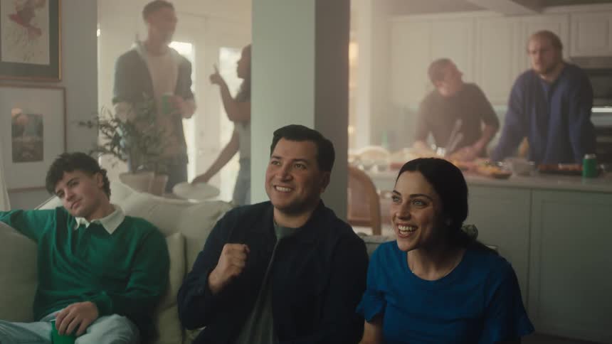 A group of friends sits on a couch in a hazy living room reacting with shock and excitement while watching a screenthey soon break into cheers and laughter highfiving and celebrating a positive moment together