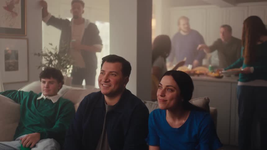 People seated on a couch smiling while watching something offscreen with others gathered in the kitchen in the background during a social gathering