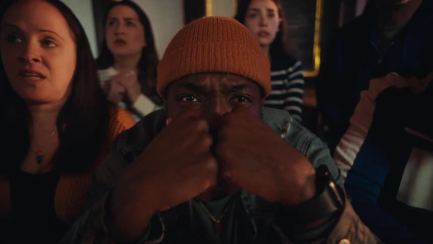 A man in an orange beanie stares intensely forward with his hands clenched near his face looking deeply anxious behind him a crowd of people watches with similarly worried and concerned expressions as if witnessing a highstakes event