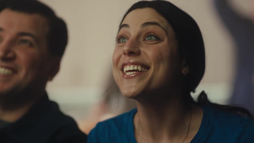 A young woman with dark hair looks up with an expression of intense excitement and then smiles broadly while watching an event with a crowd