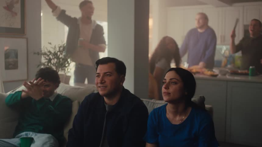 A man and a woman sit on a couch and watch a game on television while a large group of friends gathers in the background