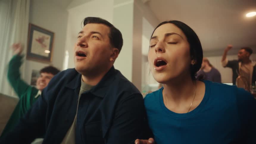 People seated on a couch reacting to an offscreen event with gestures of frustration and support in a home setting