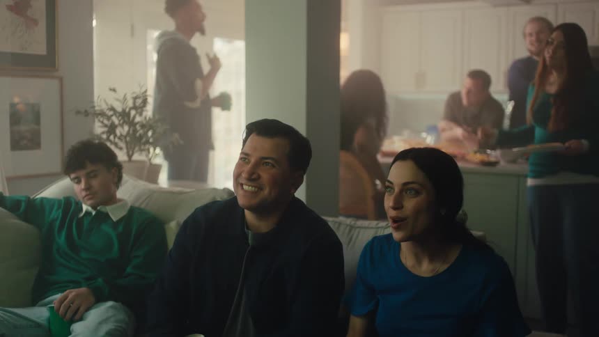 A group of friends indoors watching a game with excitement some are sitting others are standing and theyre talking as they watch