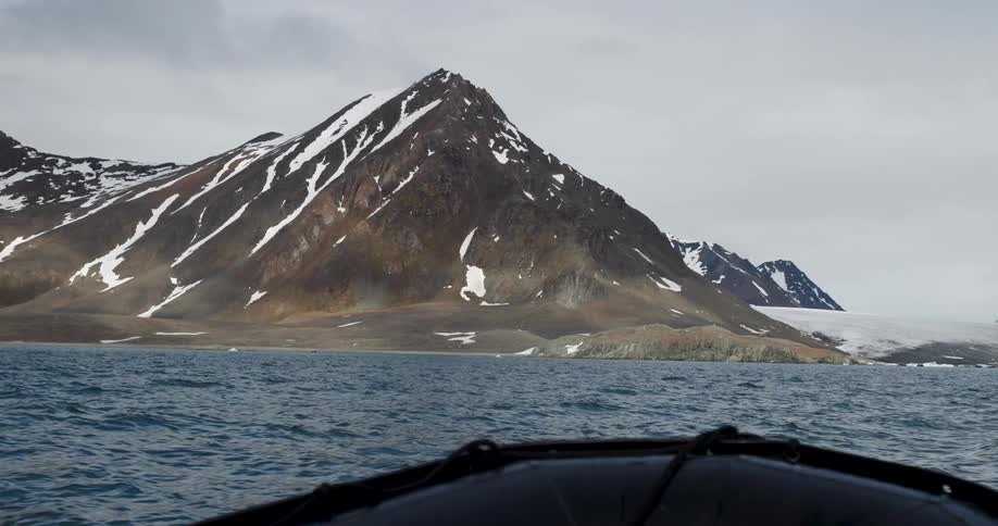 A person riding a boat across a lake toward a mountain coast with the peak covered in snow on a cloudy day