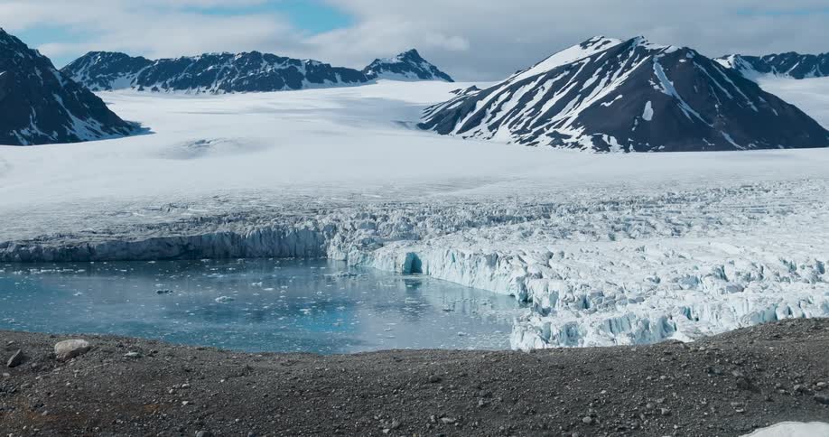 Arctic glacier calves into a cold blue meltwater pool beneath snow covered mountains