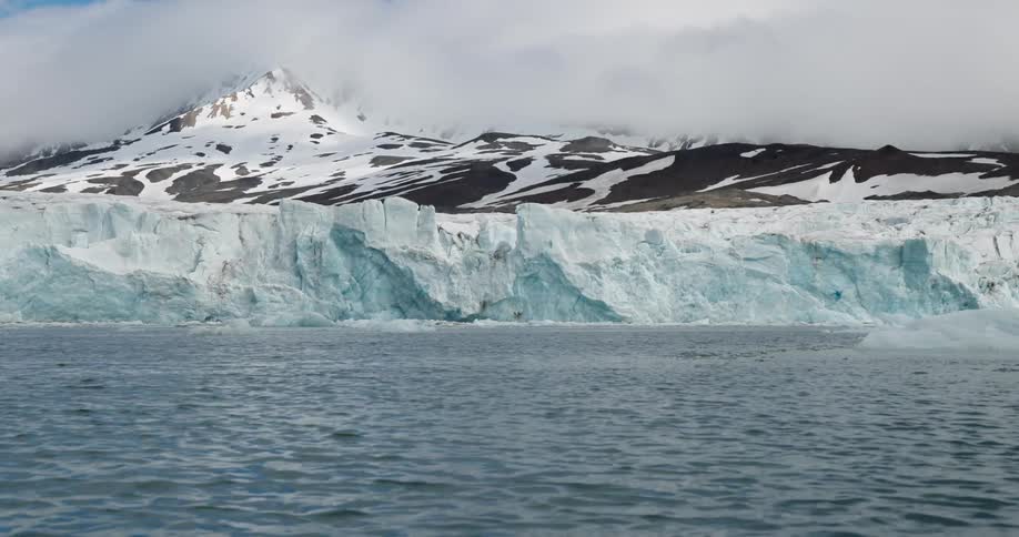 An iceberg sitting along the coastline as ocean water moves around it with a snowcovered mountain on the shore on a cloudy day