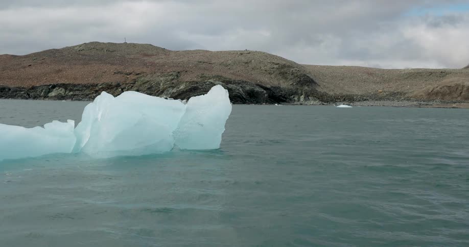 A large bright blue iceberg drifts through choppy grey waters in front of a rocky desolate shorelinethe camera pans slowly across the cold landscape capturing smaller ice fragments floating near the distant hills under an overcast sky