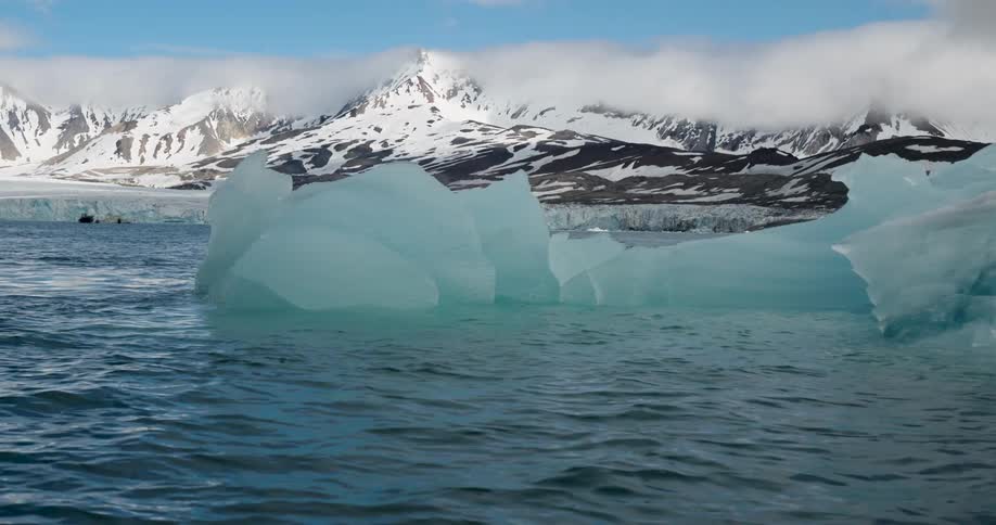 Floating ice block in blue water with snowcovered mountains in the background under a partially overcast sky
