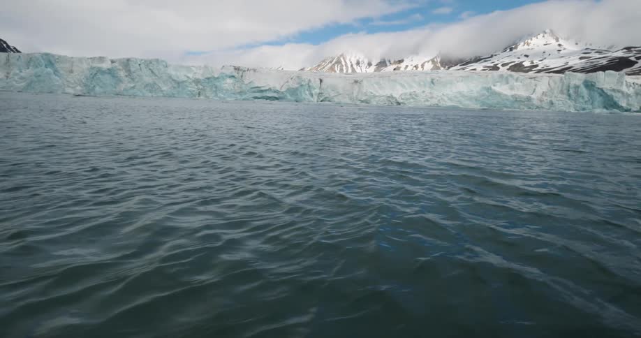 A lowangle view from the water looking toward a massive bright blue glacier wall snowy mountain peaks partially obscured by lowhanging clouds rise in the background behind the ice shelf